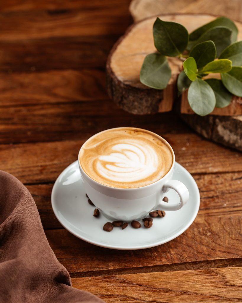 Uma xícara branca de café com leite e desenho na espuma, com grãos e uma folha em volta. A xícara está em um pires sobre uma mesa de madeira rústica.