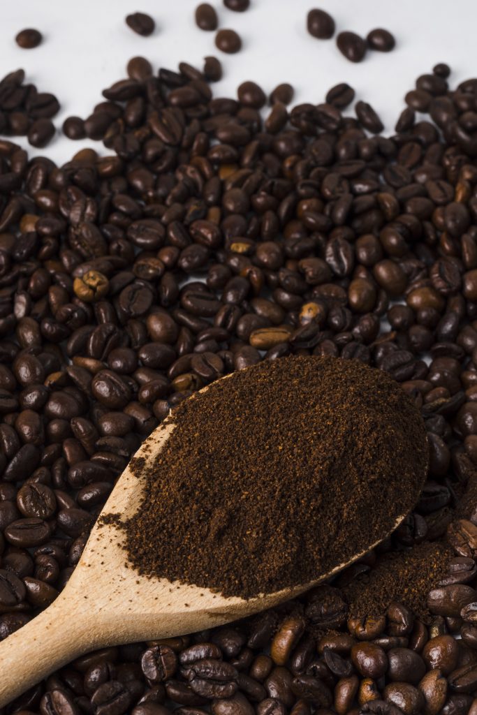 Uma colher de pau cheia de café moído, repousando sobre uma superfície coberta por grãos de café torrados.