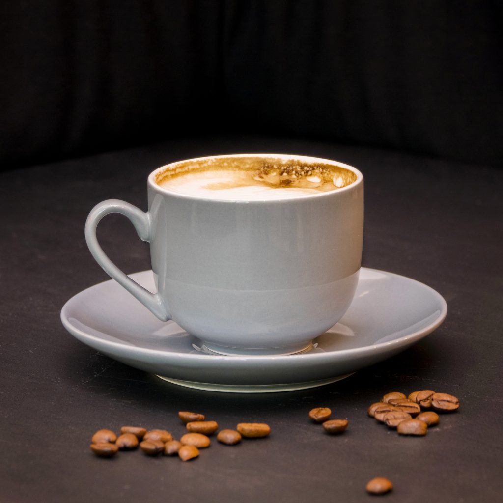 Uma xícara cinza de café com espuma cremosa, em um pires da mesma cor, com alguns grãos de café espalhados ao redor, sobre um fundo escuro.