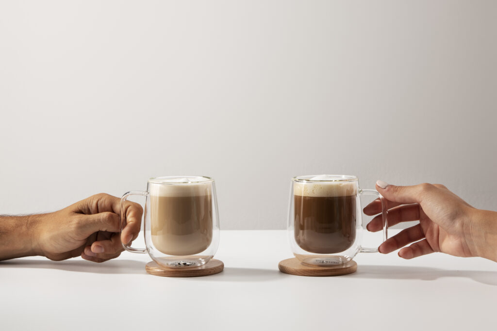 Duas mãos segurando xícaras de vidro com Café Tradicional vs. Extra Forte para comparação de cor e intensidade.