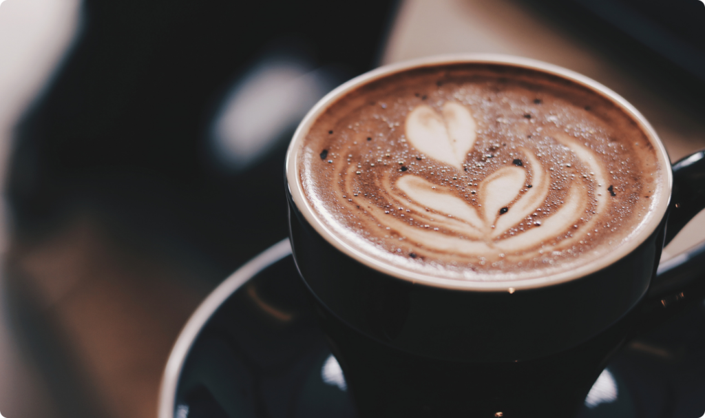 Vista superior em ângulo de uma xícara de café com arte em leite no formato de dois corações sobre uma superfície espumosa e marrom. A xícara escura está posicionada em um pires, com um fundo desfocado em tons de marrom e preto que sugere um ambiente de cafeteria.