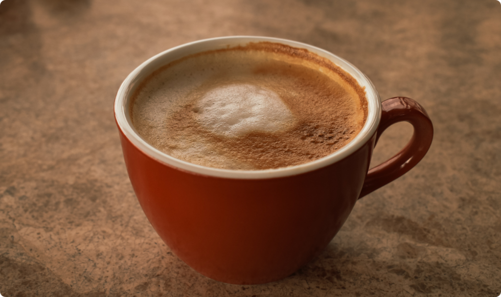 Uma xícara de cerâmica vermelha cheia de café com uma camada espessa de espuma clara e cremosa no topo. A bebida apresenta tons de marrom e bege, e a xícara está apoiada sobre uma superfície de textura marmorizada ou de pedra em tons terrosos, sob uma iluminação suave e quente.