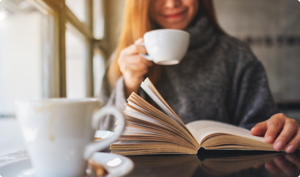 Uma mulher sorridente, em plano médio e fundo desfocado, segura uma xícara branca de café enquanto lê um livro aberto sobre uma mesa escura. Em primeiro plano, à esquerda, destaca-se outra xícara branca de café em seu pires. A cena, iluminada por uma luz natural vinda de uma janela lateral, transmite uma atmosfera de foco, produtividade e bem-estar no dia a dia.