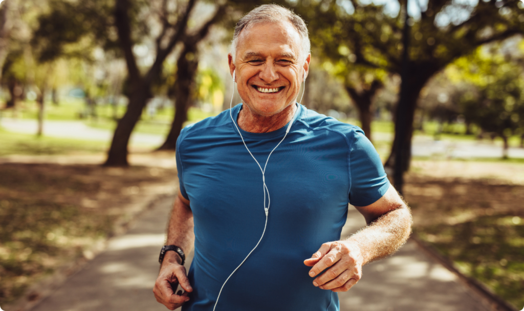 Um homem de meia-idade sorridente pratica corrida em um parque ensolarado, vestindo uma camiseta esportiva azul e fones de ouvido brancos com fio. O fundo mostra árvores e um caminho pavimentado sob luz natural, reforçando a conexão entre o consumo de café e a melhora da performance física e metabólica mencionada no planejamento.