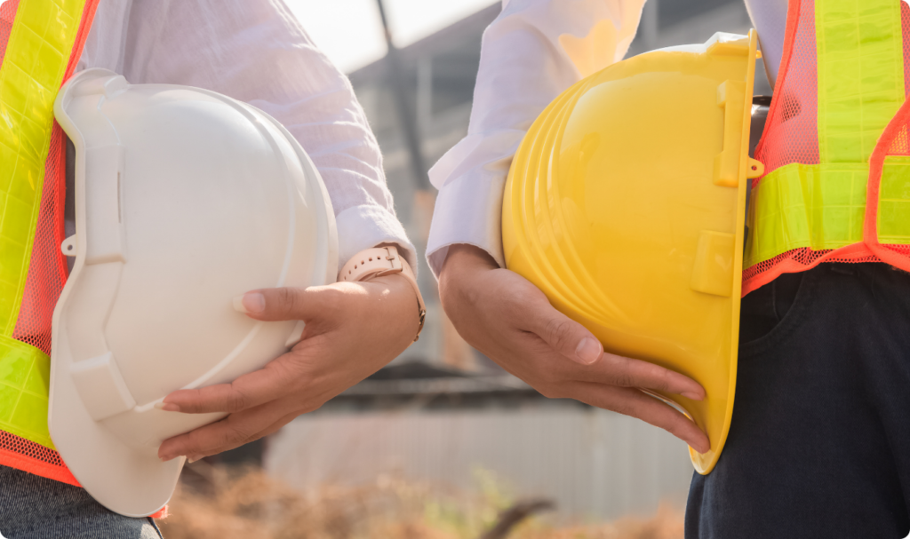 Imagem em plano detalhado de dois trabalhadores da construção civil posicionados lado a lado. Ambos vestem coletes refletivos laranja com faixas amarelas e seguram seus capacetes de segurança (um branco e um amarelo) sob os braços. A imagem ilustra o público de trabalho braçal que busca no café o rendimento necessário para rotinas exaustivas.